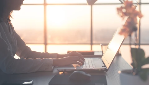 Woman working on laptop at desk by sunlit window during golden hour