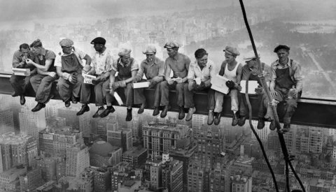 Workers perched on steel beams high above a cityscape during the construction of a skyscraper.