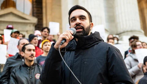 Man with dark beard speaking into microphone at outdoor public gathering with crowd in background