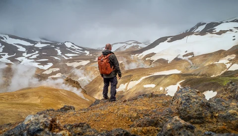 Hiker in orange backpack standing on rocky outcrop overlooking geothermal landscape with snow-capped mountains