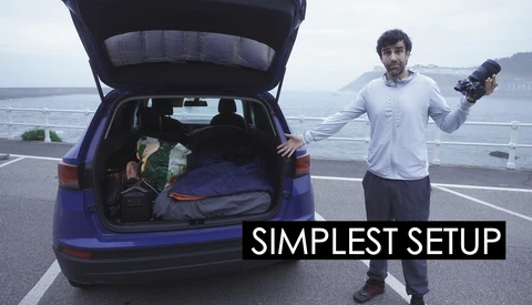 Filmmaker holding camera beside open car trunk packed with gear overlooking coastal highway