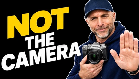 Man in navy cap and jacket holding a Fujifilm camera with palm raised, with yellow and white text reading 'NOT THE CAMERA'
