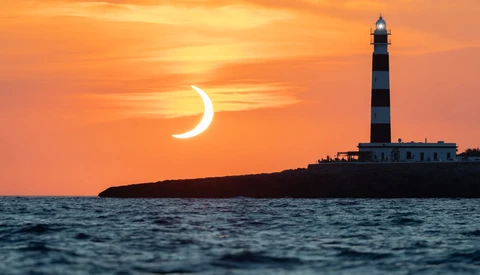 Crescent moon silhouetted against orange sunset with striped lighthouse on rocky island