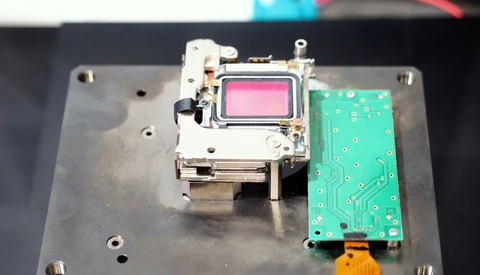 Disassembled camera internals on a metal plate: a metal-framed module with a pink-tinted sensor beside a green circuit board