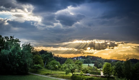 Sun rays break through dark storm clouds over a rolling green landscape of trees and a winding path at sunset.