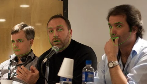Three men on a panel, center speaker talking into a microphone, green face-tracking outlines overlaid on their faces.