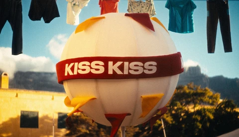 Oversized white balloon with red 'KISS KISS' band floating above landscape with clothesline of garments