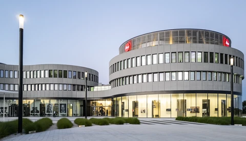 Curved modern building at dusk with red Leica logo signs on its upper level and an illuminated glass ground floor.