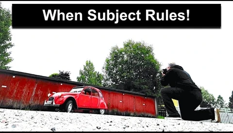 Red vintage Citroën 2CV parked against red corrugated metal wall with large dark sculptural figure looming above
