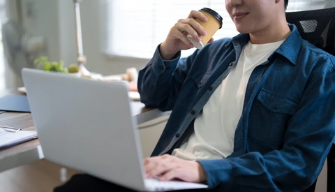 Person holding coffee cup while working on laptop at desk