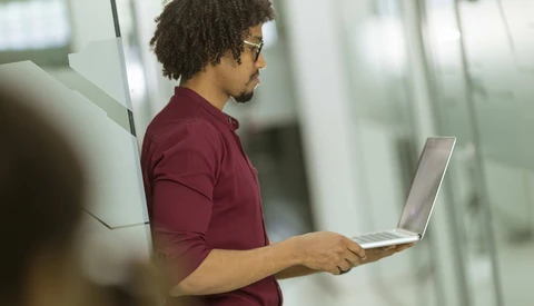 Man in maroon polo shirt holding open laptop, standing in modern office corridor with glass partitions