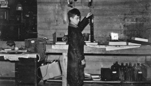 Woman reaching upward in a laboratory or studio workspace with equipment and bottles visible