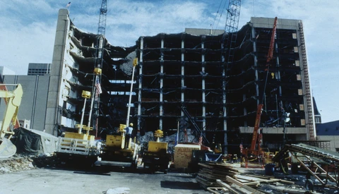 Partially collapsed multistory concrete building with exposed floors, surrounded by cranes, debris and yellow machinery.