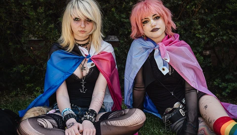 Two cosplayers in coordinated black outfits with flowing capes sitting on grass against greenery