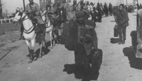 Historical black and white photograph of a military procession with mounted officer on white horse leading infantry troops down a street