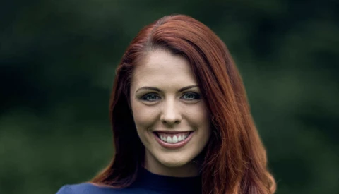 Woman with long reddish-brown hair smiling at camera against blurred green background