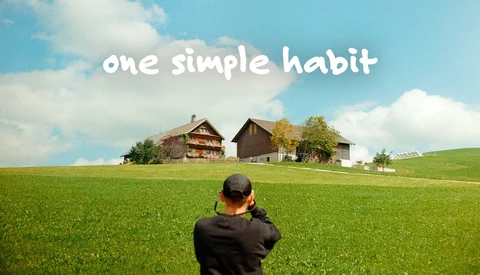 Person photographing rural farmhouses across an open green field under blue sky with text overlay