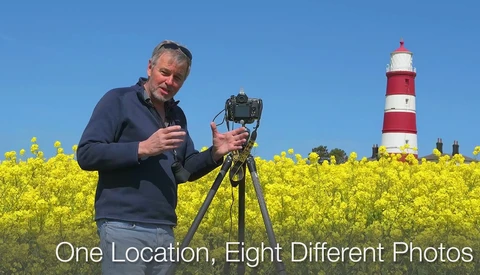 Photographer holding camera on tripod in yellow rapeseed field with red and white striped lighthouse in background