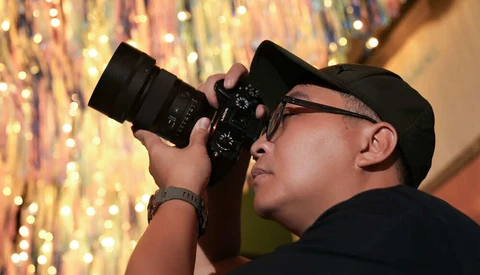 Photographer in black cap and glasses holding DSLR camera, shooting toward warm-toned bokeh backdrop