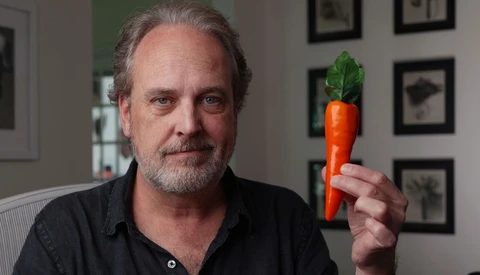 Man holding a fresh carrot with green leafy top in an indoor studio setting