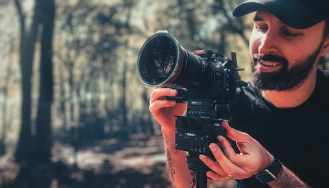 Bearded cinematographer in cap holding cinema camera with telephoto lens in forest