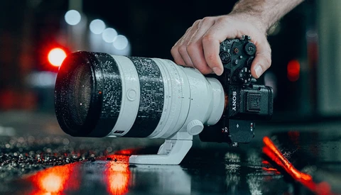 Photographer's hand holding Sony mirrorless camera with telephoto lens on wet surface with red bokeh lights