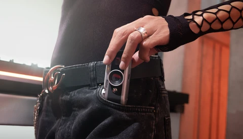 Person wearing black clothing and mesh sleeve holding a compact camera at hip level