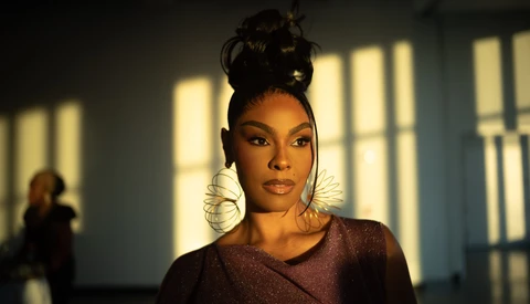 Woman with hair in a high bun and sculptural wire hoop earrings, lit by warm sunlight streaming through windows onto a wall behind her.