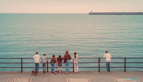 Group of people standing at a seaside promenade railing, overlooking turquoise water with a cargo ship on the horizon