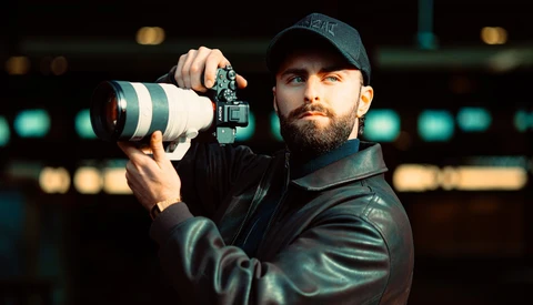 Bearded photographer holding a telephoto lens and mirrorless camera against blurred stadium lights