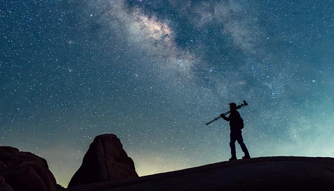 Silhouette of a person carrying a tripod over one shoulder, standing on a rock ridge beneath a bright Milky Way sky.