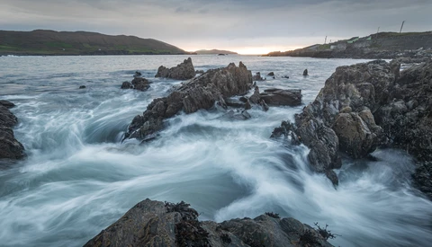 Long exposure of silky surf flowing around dark, jagged rocks on a coastline under a moody overcast sky