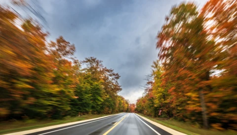 Blurred motion shot of a two-lane road lined with autumn trees in orange and red foliage under a gray, overcast sky.