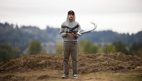 Man in a gray hoodie and cap holding a video camera rig with a top-mounted shotgun microphone on a dirt mound outdoors