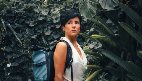 Woman with short dark hair in a white draped top and teal backpack, standing among lush tropical leaves