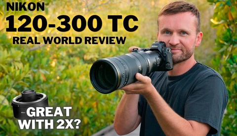 Bearded man in a dark t-shirt raising a Nikon Z camera with a telephoto zoom lens outdoors among green foliage.