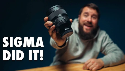 Bearded man holding a Sigma camera lens toward the camera against a dark blue background
