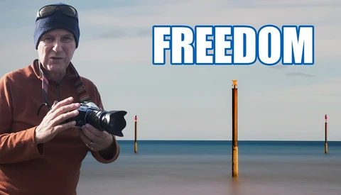 Photographer holding a camera on a beach with wooden pilings and calm water, with text overlay reading 'FREEDOM'