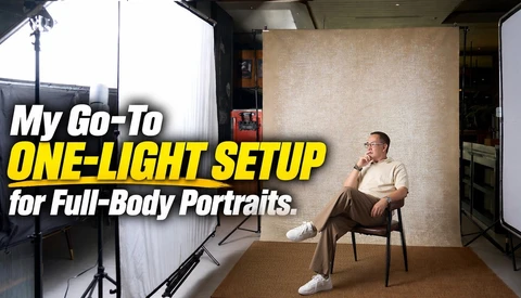 Man seated on a wooden chair in a studio, posed against a textured beige backdrop with a softbox on a stand at left.