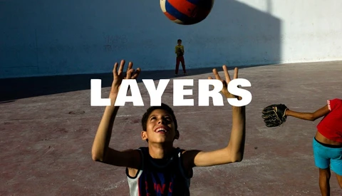 Smiling boy with raised arms looking up at a ball on a sunlit concrete court, with a shadowed wall behind him