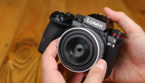 Hands holding a Panasonic Lumix mirrorless camera with a compact silver-rimmed pancake lens facing the viewer