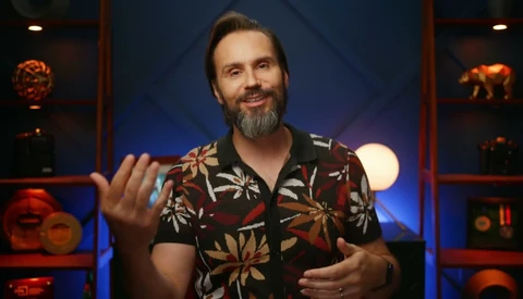 Smiling bearded man in a floral short-sleeve shirt gesturing with both hands in front of a blue-lit shelf set.
