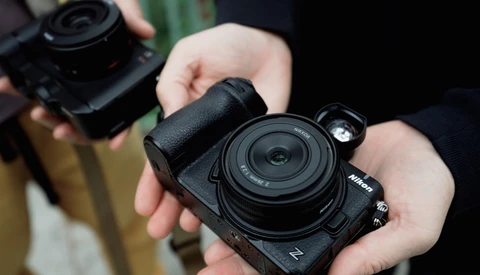 Hands holding a black Nikon Z-series mirrorless camera with a compact pancake lens, another camera blurred in the background