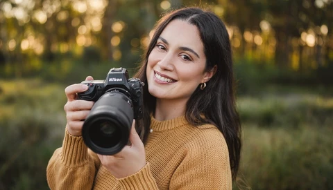 Smiling woman with long dark hair in a mustard sweater holding a Nikon mirrorless camera with a large zoom lens, outdoors in soft light.
