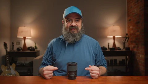 Bearded man in a blue cap and blue shirt pointing at a black camera lens standing on a wooden table