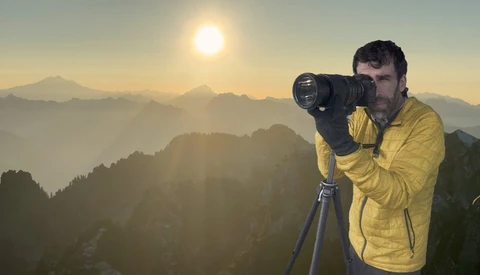 Photographer in a yellow jacket and gloves shooting through a telephoto lens on a tripod above layered mountain ridges at sunrise