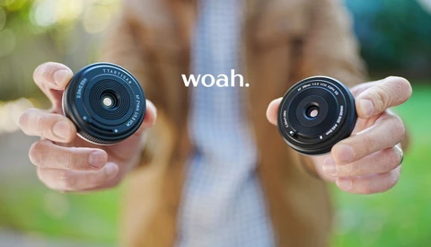 Person holding two compact camera lenses toward the viewer, with the word 'woah.' overlaid on a blurred outdoor background