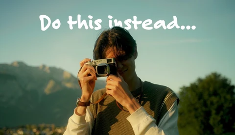 Person in a knit vest raising a silver compact film camera to their eye, with sunlit mountains and trees behind them.