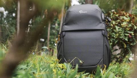 Black Lowepro camera backpack with orange zipper pulls resting on grass in a tropical garden.