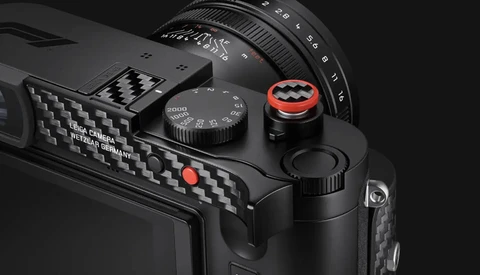Close-up product shot of a black Leica camera's top plate and carbon-fiber grip, with a red-ringed shutter release and speed dial.